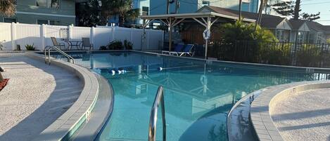 A swimming pool with a tiled deck, lounge chairs, and a covered area with a roof.