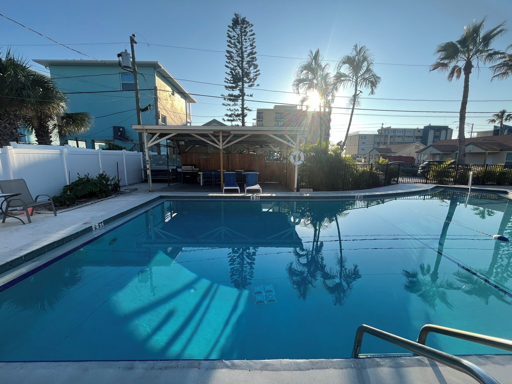 Una piscina con sillas de descanso y un área de estar cubierta, rodeada de palmeras y edificios.