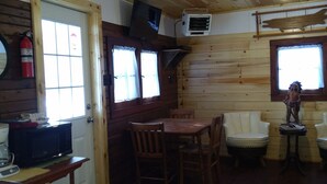 A cozy cabin interior with wooden walls, a dining area with chairs and a table, a television mounted on the wall, and a fire extinguisher near the door. - Cabin #5 On Beautiful Brevort Lake (Moran)