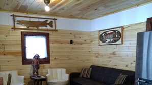 A cabin interior with wooden walls, a wooden ceiling, a wooden fish decoration, a framed picture, a sofa, a chair, a small table, and a refrigerator. - Cabin #5 On Beautiful Brevort Lake (Moran)