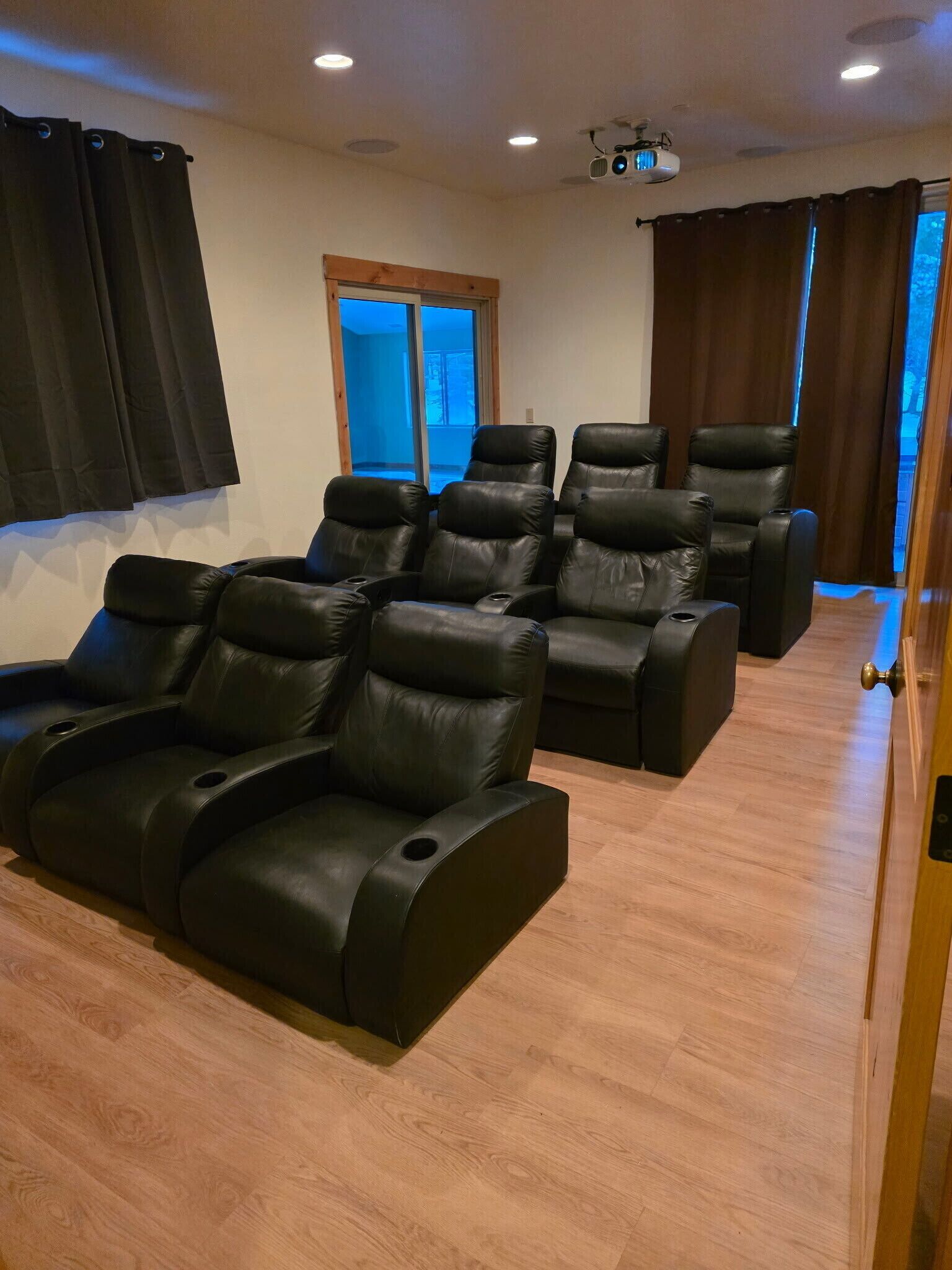 Una sala con varias sillas reclinables de cuero negro dispuestas en filas, piso de madera y proyector en el techo.