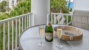 Une bouteille de champagne KORREL, deux verres et un plateau en bois sur une table en verre, avec une balustrade blanche et une vue sur des palmiers et des immeubles.