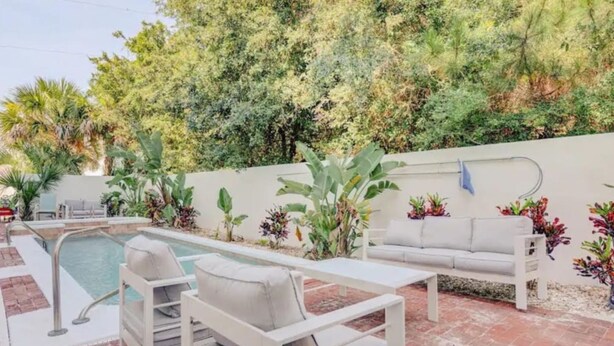 A pool area with a white sofa and chairs, surrounded by lush greenery and a white wall. - Ocean Song - Hot Tub (St. Augustine)