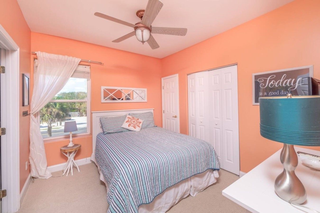 Quarto com cama, ventoinha no teto, janela com cortinas, mesa de cabeceira e um enfeite na parede.