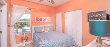 Quarto com cama, ventoinha no teto, janela com cortinas, mesa de cabeceira e um enfeite na parede.
