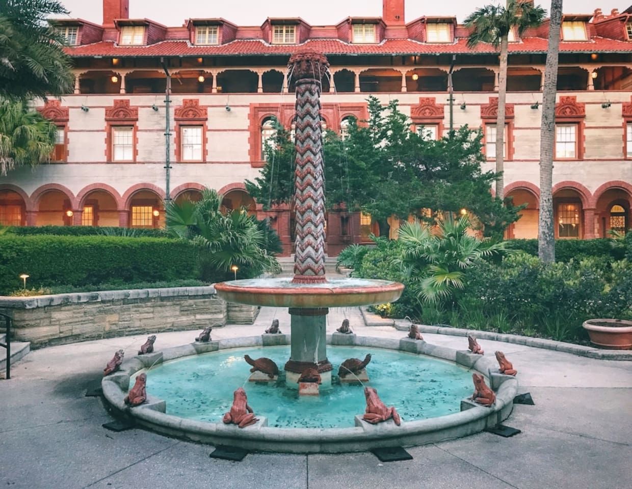 A fountain with a central column and frog statues, surrounded by a circular pool, with a building in the background.