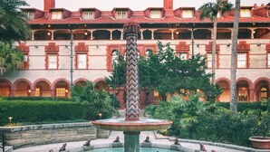 A fountain with a central column and frog statues, surrounded by a circular pool, with a building in the background. - Ocean Song - Hot Tub (St. Augustine)