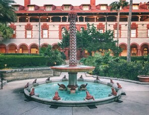 A fountain with a central column and frog statues, surrounded by a circular pool, with a building in the background. - Ocean Song - Hot Tub (St. Augustine)