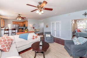 A spacious living area with a white sofa, a grey armchair, and a coffee table. There are ceiling fans, a kitchen in the background, and a door leading to another room.