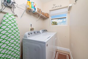 Een wasserette met een voorlader wasmachine en droger, een droogrek met schoonmaakmiddelen, een raam met uitzicht en een wandplank met manden.