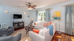 A living room with a ceiling fan, a television, a sofa set, a coffee table, and a painting on the wall. - Ocean Song - Hot Tub (St. Augustine)