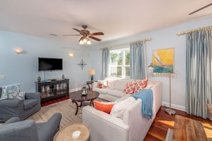 A living room with a ceiling fan, a television, a sofa set, a coffee table, and a painting on the wall. - Ocean Song - Hot Tub (St. Augustine)