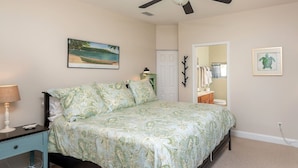 A bedroom with a bed, a nightstand, a ceiling fan, a painting, a mirror, and a door leading to another room. - Ocean Song - Hot Tub (St. Augustine)