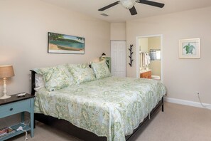A bedroom with a bed, a nightstand, a ceiling fan, a painting, a mirror, and a door leading to another room. - Ocean Song - Hot Tub (St. Augustine)