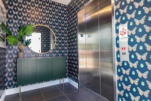 Un ascensor con detalles metálicos, una pared con estampado de mariposas, un espejo redondo y un radiador verde.