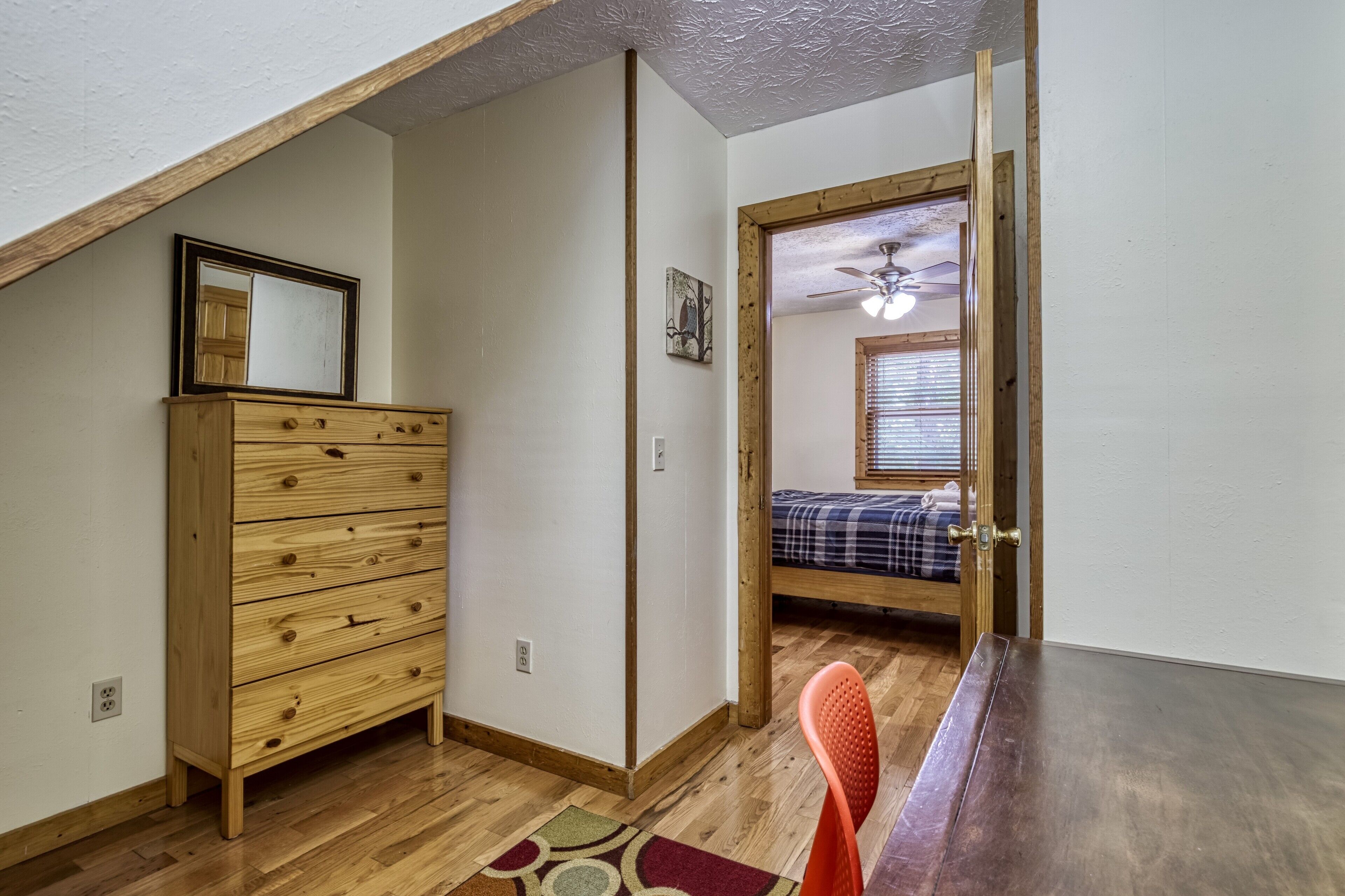 A bedroom with a wooden dresser, a bed with a plaid blanket, a ceiling fan, and a window with blinds.
