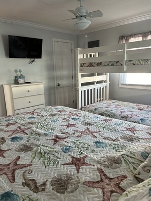 Une chambre avec deux lits, une commode, un téléviseur, un ventilateur de plafond et une fenêtre avec des rideaux.