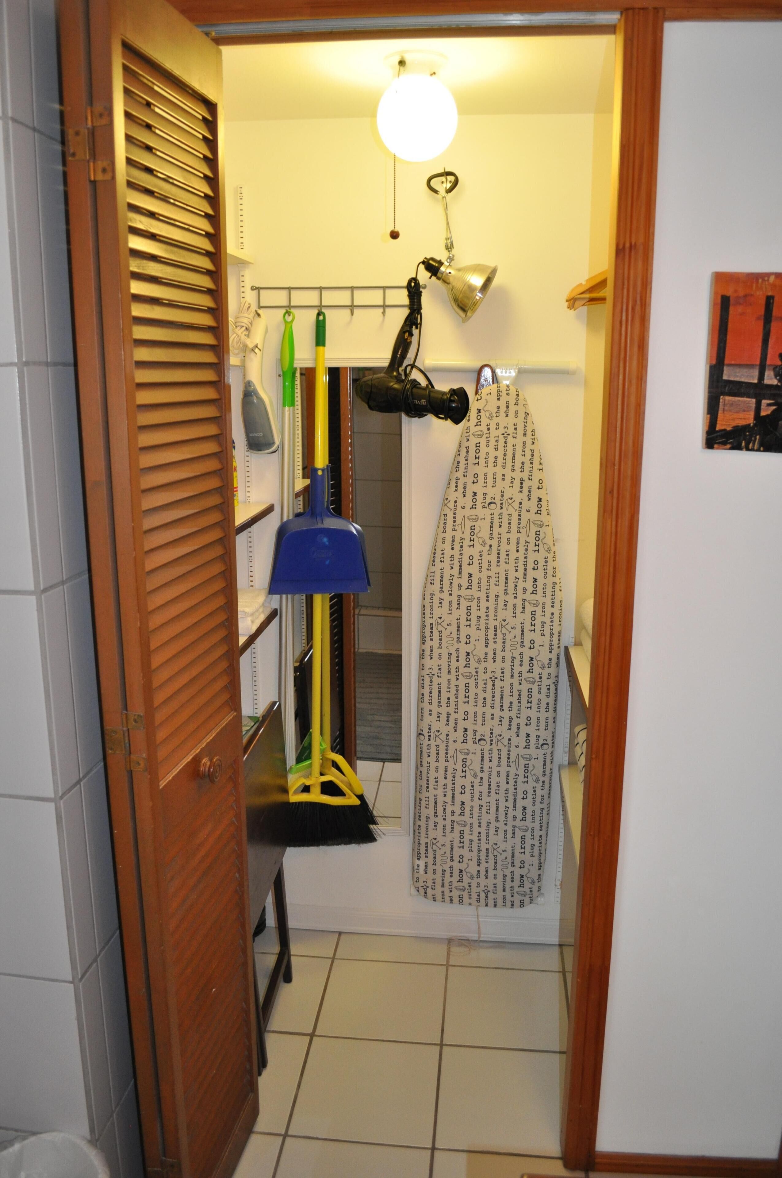 A narrow hallway with a wooden door, a hanging lamp, a rack with cleaning supplies, and a patterned shower curtain.