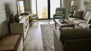 Un salon comprenant un canapé, une table basse, un téléviseur et un fauteuil. Il y a une fenêtre avec des rideaux et une vue sur l’extérieur.