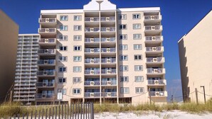 Un hôtel à plusieurs étages doté de balcons, avec un rez-de-chaussée dont le sol est recouvert de sable, et un ciel d’un bleu limpide.