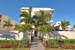Un hotel moderno de varios pisos con entrada central, rodeado de palmeras y un jardín ajardinado.