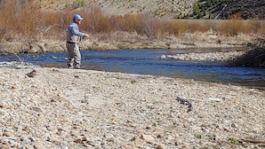 A man fishing by a river with a rocky shore and forested hills in the background. - Riverhouse Retreat on East Fork of the Bitterroot - Stunning Views & Tranquility (Conner)