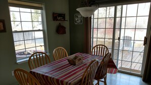 A dining area with a table set for six, wooden chairs, a striped tablecloth, and a view of the backyard through large windows. - Riverhouse Retreat on East Fork of the Bitterroot - Stunning Views & Tranquility (Conner)