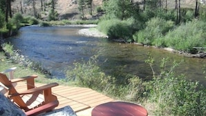 A wooden deck with a table and chairs overlooking a river, surrounded by greenery and trees. - Riverhouse Retreat on East Fork of the Bitterroot - Stunning Views & Tranquility (Conner)