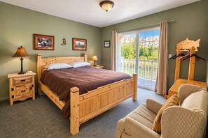 A bedroom with a wooden bed, a nightstand, a lamp, a chair, a balcony with a view of greenery, and a coat rack. - Riverhouse Retreat on East Fork of the Bitterroot - Stunning Views & Tranquility (Conner)