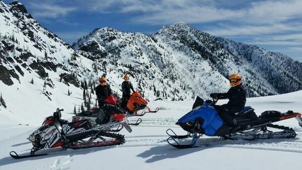 A snow-covered mountain landscape with snowmobiles and riders.
