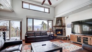 A spacious living room with a stone fireplace, leather sofas, a large TV, and a view of the mountains through large windows. - Ski In/Out-Sleeps 18, 9,000 Sq Ft-Hot Tub-Pool Table-Mtn Views-Theatre Rm (Breckenridge)