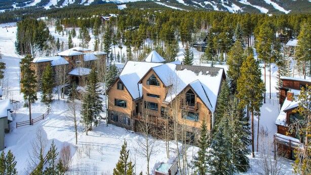 A large, two-story lodge with a prominent central chimney, surrounded by snow-covered trees and mountains in the background. - Ski In/Out-Sleeps 18, 9,000 Sq Ft-Hot Tub-Pool Table-Mtn Views-Theatre Rm (Breckenridge)