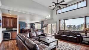 A spacious living room with a large sectional sofa, a coffee table, and a fireplace. There is a kitchen area with wooden cabinets and a large window overlooking a scenic view. - Ski In/Out-Sleeps 18, 9,000 Sq Ft-Hot Tub-Pool Table-Mtn Views-Theatre Rm (Breckenridge)