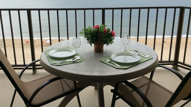A round table with a green placemat, two white plates, a glass, and cutlery on a balcony overlooking the sea.