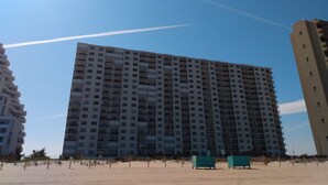 A high-rise building with multiple floors, a sandy ground, and a clear sky.