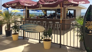 A beachside bar with maroon umbrellas, a sign prohibiting food, beverages, or alcohol on the deck, and a view of the ocean.