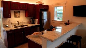 A kitchen with dark wooden cabinets, a stainless steel refrigerator, a granite countertop, and a window providing a view of greenery outside. - End Unit Yards from Ocean Looking East With Major Sunrises (Kapaa)