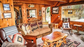 Interior acogedor de una cabaña con un sofá de madera, una mesa de madera y un sillón mecedor. Se observan cuadros enmarcados en las paredes y una ventana con vistas al exterior.