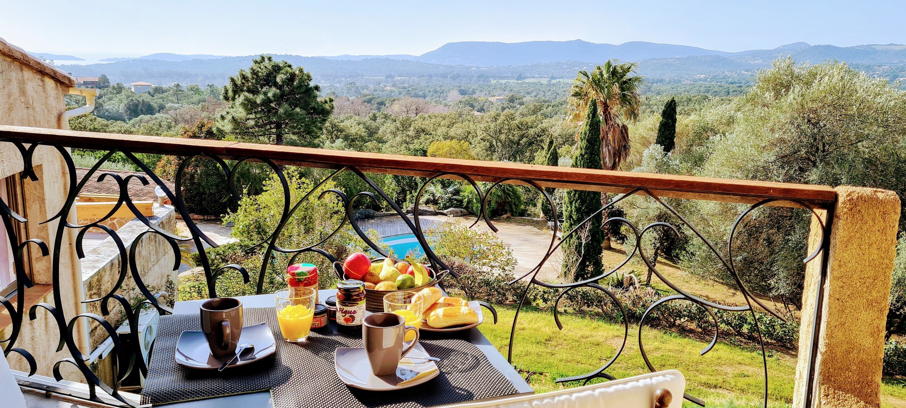 Un balcon avec une table dressée pour le petit-déjeuner, offrant une vue panoramique sur un paysage magnifique, avec des arbres et des montagnes.