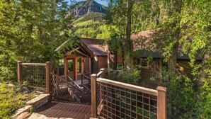 A rustic cabin with a wooden staircase leading to a porch, surrounded by lush greenery and a mountainous backdrop. - Telluride Riverfront House with Mountain Views (Telluride)