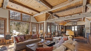 A spacious living area with a high wooden ceiling, large windows offering a view of a snowy landscape, and a cozy seating arrangement. - Telluride Riverfront House with Mountain Views (Telluride)
