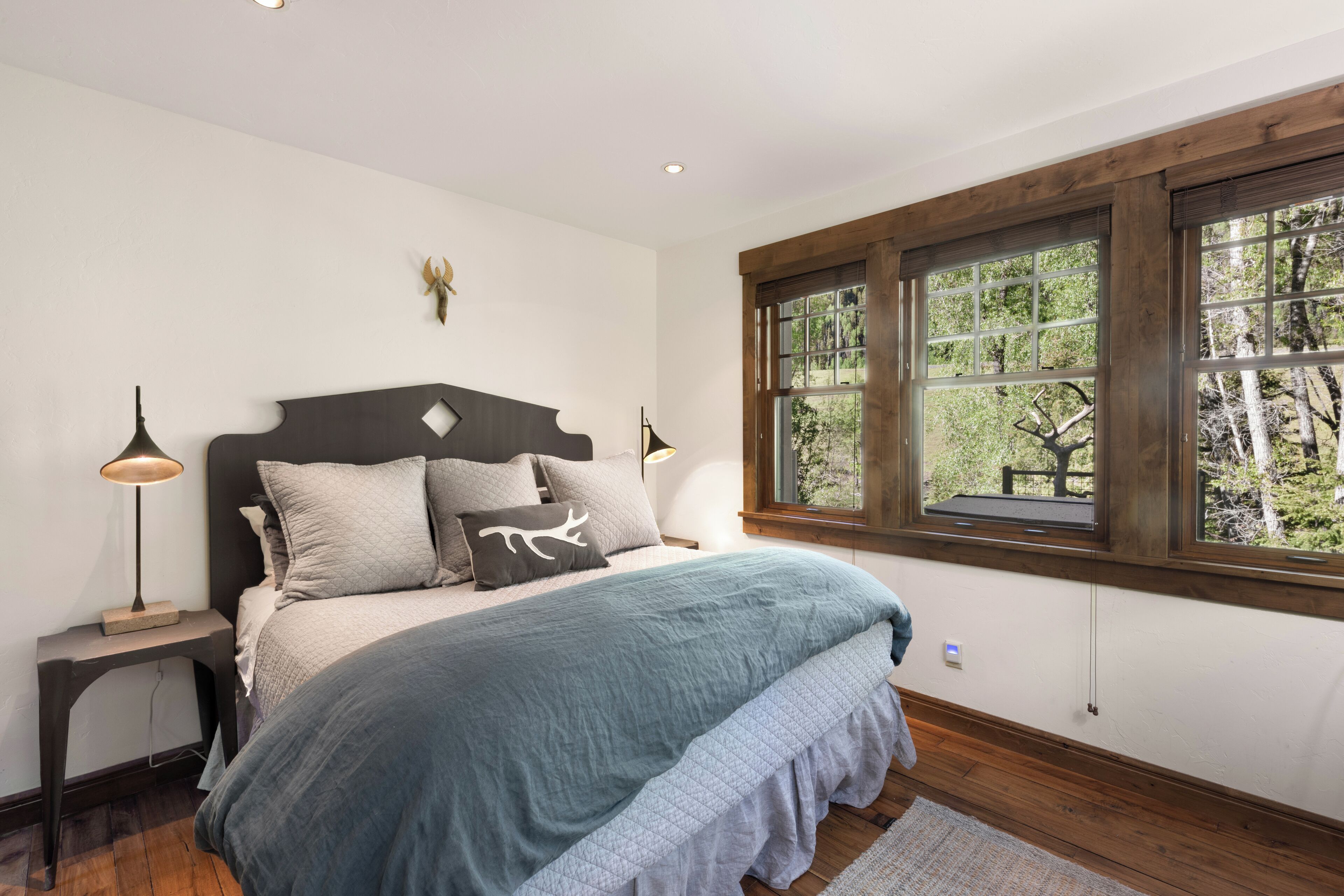 Une chambre avec un grand lit, une tête de lit en bois, deux lampes de chevet, une fenêtre donnant sur les arbres et un luminaire fixé au mur.