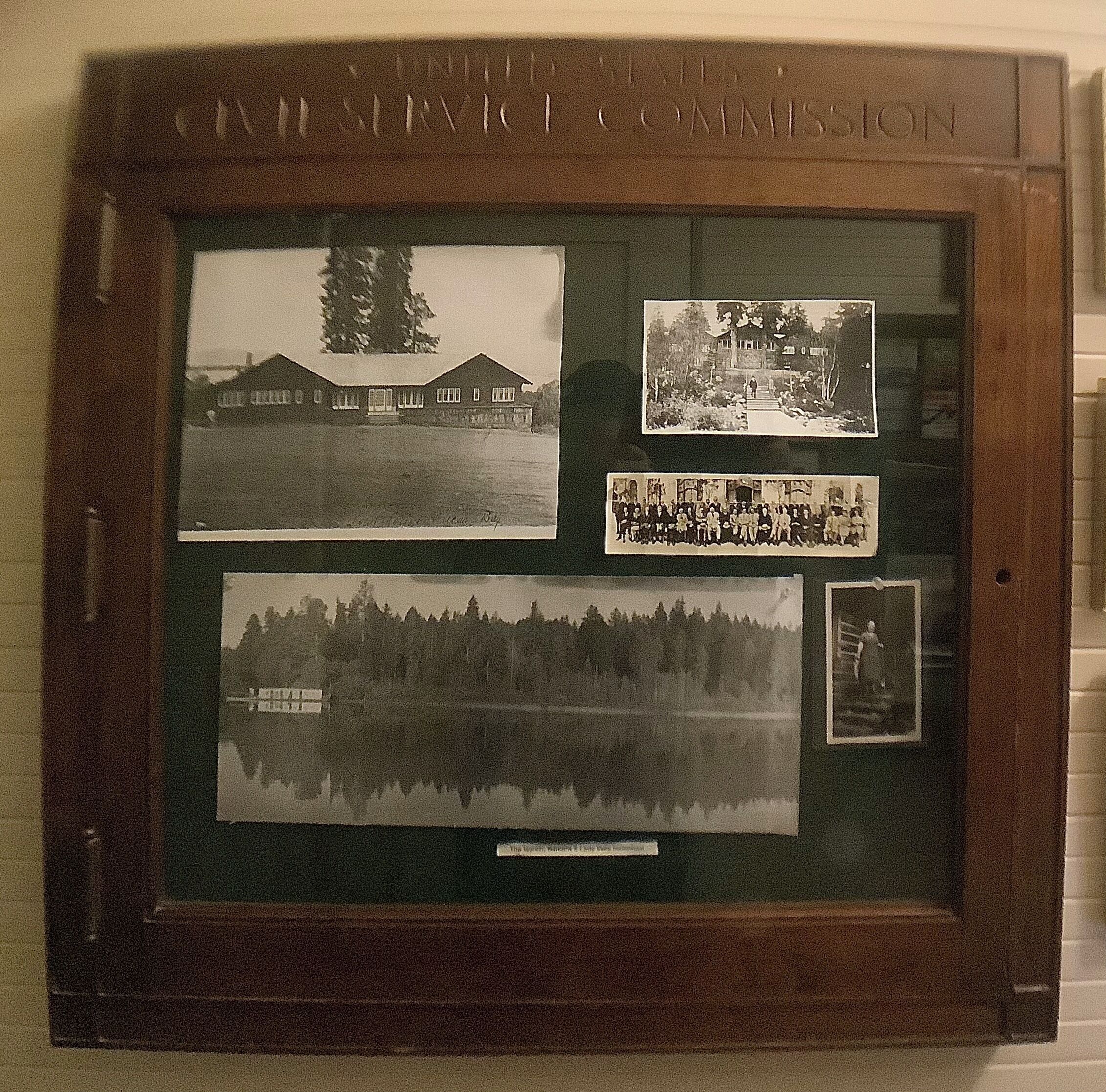 A framed display with four black and white photographs, including a building, a group of people, a forest, and a lake scene.