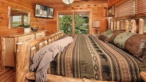 Un dormitorio de cabaña de madera con cama, ventilador de techo, televisor y ventana con vistas a follaje.