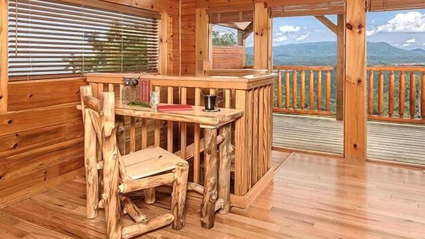 Una terraza de madera con una mesa y silla de madera rústica, un estante con objetos decorativos y vistas a montañas a través de la barandilla.