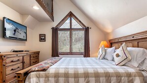 A bedroom with a large bed, a wooden dresser, a window with curtains, and a television on the wall.