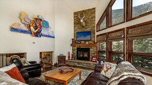 A cozy living room with a fireplace, leather sofas, a coffee table, and a large window with a view of trees.