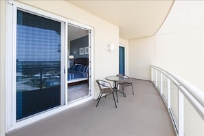 Un balcon doté d’une porte vitrée donnant sur une chambre à coucher où se trouvent un lit et une chaise. On y trouve également une petite table entourée de chaises.