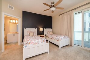 Une chambre avec deux lits blancs, un ventilateur de plafond, une table de chevet, une lampe, une salle de bain avec un meuble-lavabo et des luminaires, et une grande fenêtre avec des rideaux.