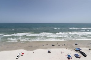 Une plage avec une voiture blanche garée sur le sable, plusieurs parasols de plage et quelques personnes qui se promènent.